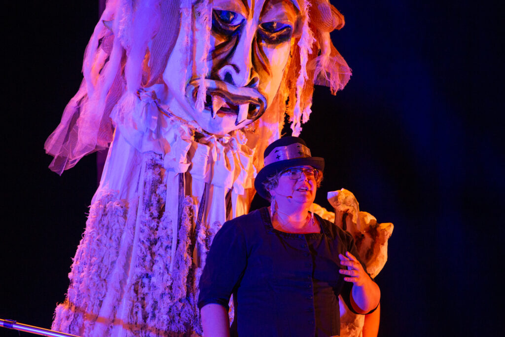 Photo of Jennifer Pictou in a black dress and black hat, standing in front of the puppet Jenu the ice giant