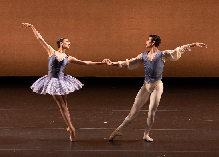 Photo of two dancers, one in a light tutu up on pointe shoes, the other in light tights and a blue top supporting her in a duet