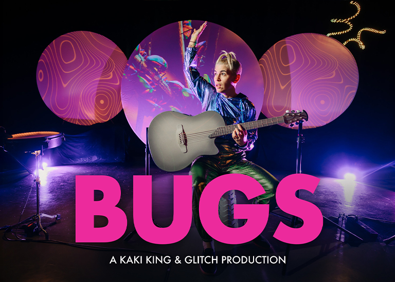 Photo of Kaki King sitting on a stool playing guitar in front of 3 large circles, that have a projected green color on them.
