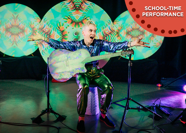 Photo of Kaki King sitting on a stool playing guitar in front of 3 large circles, that have a projected green color on them.
