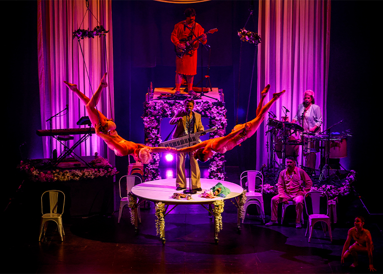 Photo of two performers suspended in the air on trapeze bars, they are swinging over a stage featuring a wedding band