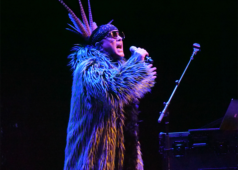 Photo of Firefly singing on stage wearing an Indigenous head dress, sunglasses and fur coat