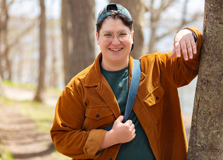 Photo of Lindz Amer wearing an orange jacket, standing next to a tree.