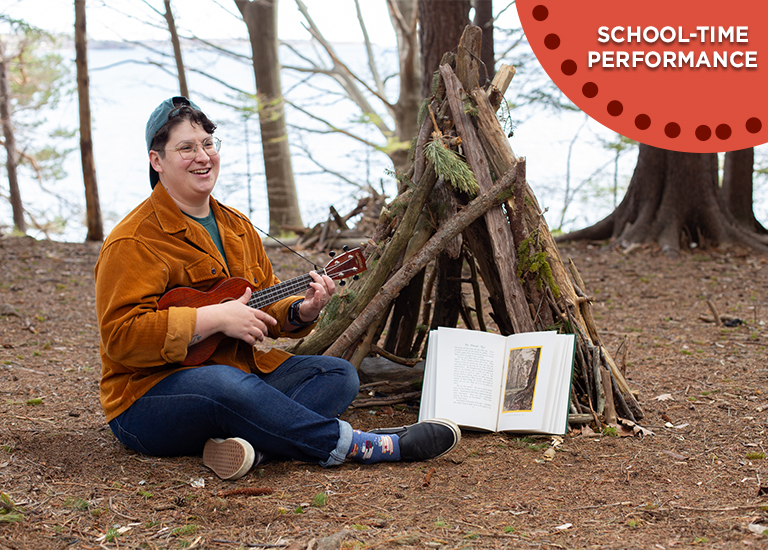 Photo of Lindz Amer wearing an orange jacket, sitting on the ground next to a stick fairy house, playing a ukulele.