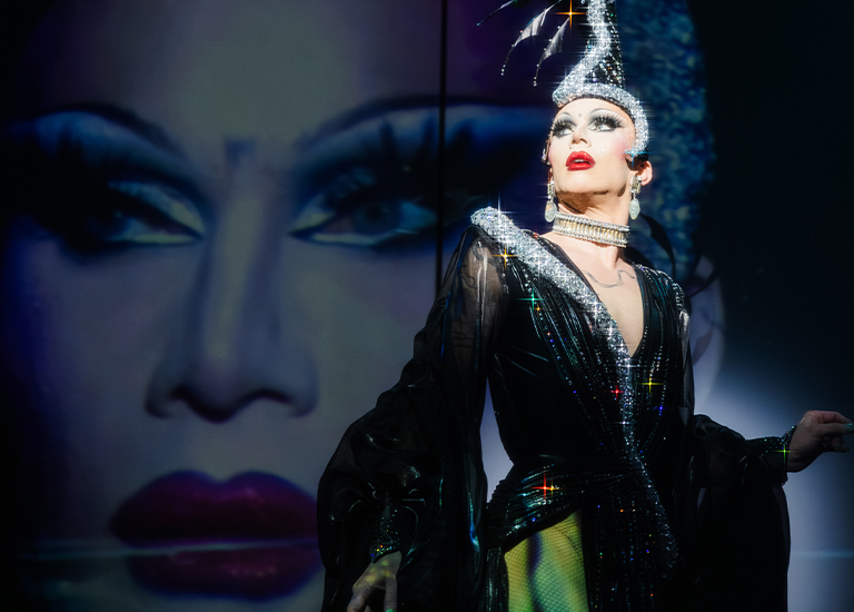Photo of Sasha Velour in long black dress, and a tall head dress standing in front of a projected image of her face