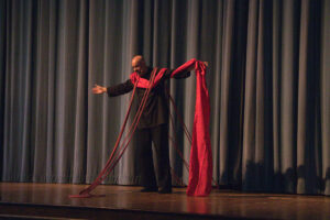 Photo of Antonio Rocha wearing a black shirt and pants, swaying on stage like a ships mast in the sea, draped with long red cloth like a scarf.