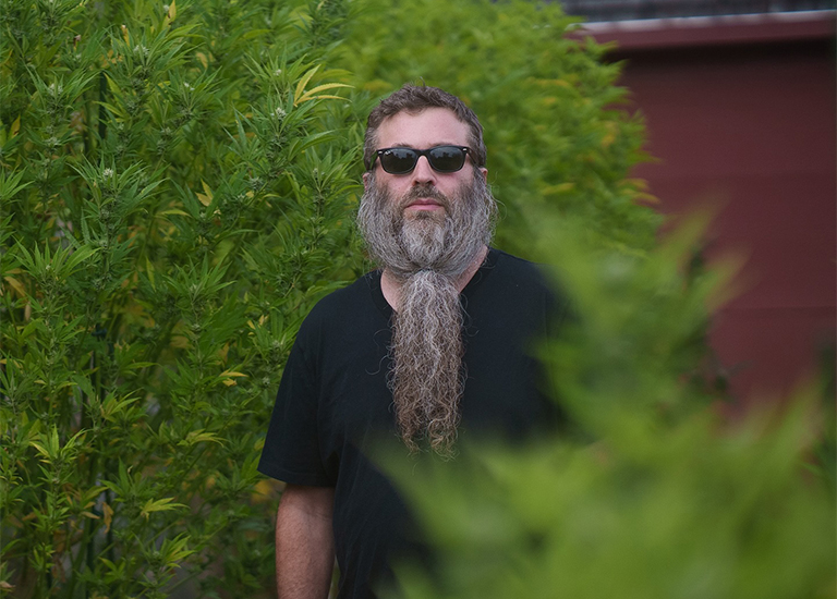 Photo of Jamie Saft wearing a pair of dark sunglasses, standing in front of a green shrub.